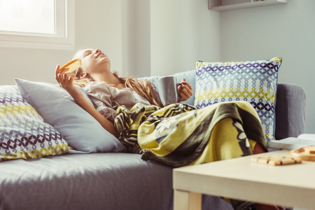 10 years old pre teen girl wrapped in blanket relaxing with cocoa and breakfast on a couch in living room. Comfortable weekend morning.の写真素材