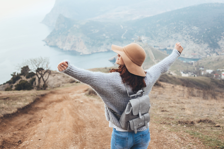Girl wearing hat and sweater travel in mountains alone. Cold weather, calm scene. Backpacker walking outdoors in fall, back view over landscape. Wanderlust photo series.の写真素材