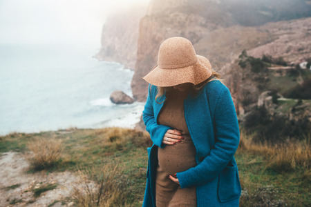 Pregnant girl wearing coat and hat traveling in mountains. Cold weather, calm scene. Happy and healthy maternity. Wanderlust photo series.の写真素材