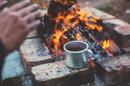 Traveler warming his hands by the campfire outdoors. Tea or coffee in aluminum mug on background. Camping detail, travel lifestyle photo.の写真素材