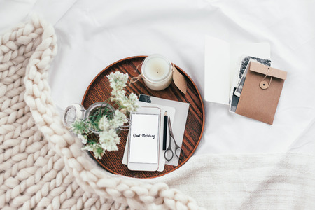 Top view of wooden tray with smartphone, old photos, candle and spring flowers on white bedding. Relaxing and posting to blog in bed at home.の写真素材