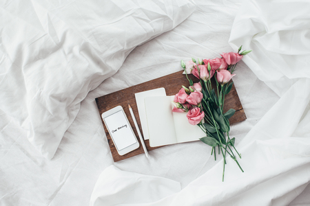Wooden tray with paper sketchbook, smartphone and spring flowers on clean white bedding. Good morning concept.の写真素材