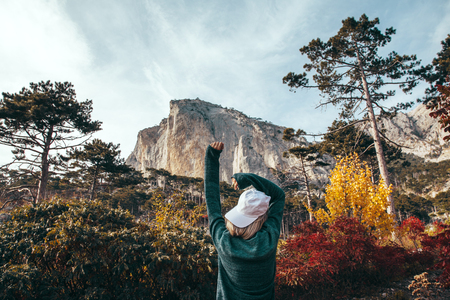 Back view of traveler hipster girl wearing cap and sweater standing alone in autumn woods and looking at mountain hill. Cold weather, fall colors. Wanderlust concept.の写真素材