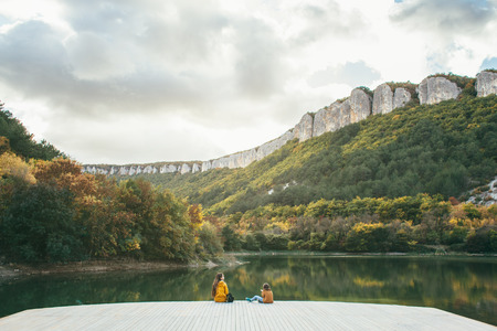 Children spending time by the lake in autumn. Two kids sitting on wooden pier and looking at the landscape. Fall weekend in the open air.の写真素材