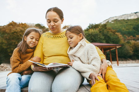 Family spending time together outdoors in autumn. Mom with her daughters reading book in park. Fall weekend in the open air.の写真素材