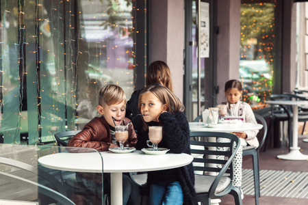 Children having fun in outdoor cafe. Kids talking and drinking cocoa for the breakfast in the city street in autumn.の写真素材