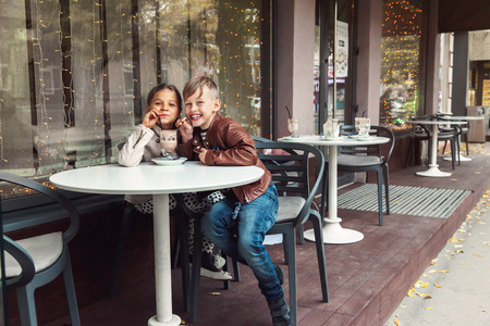 Children having fun in outdoor cafe. Kids talking and drinking cocoa for the breakfast in the city street in autumn.の写真素材