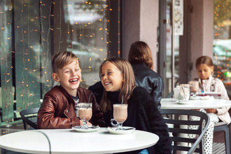 Children having fun in outdoor cafe. Kids talking and drinking cocoa for the breakfast in the city street in autumn.の写真素材