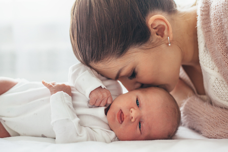Home portrait of a newborn baby with mother on the bed. Mom kissing her child.の写真素材