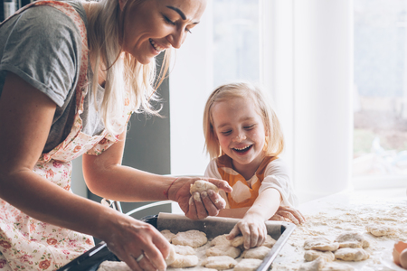 Beautiful blond mom teaching her daughter cooking on the kitchen. Parent making everyday breakfast together with child. Family at home lifestyle photo.の写真素材
