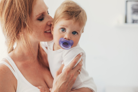 Portrait of beautiful loving mom holding on hands and kissing her 8 months old baby with nipple in bedroomの写真素材