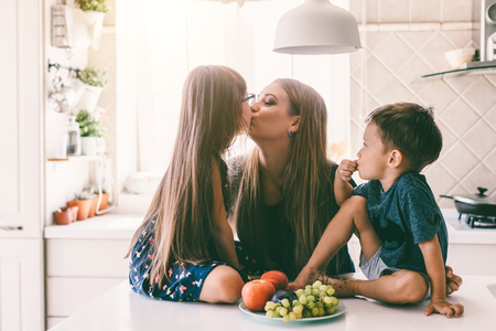 Mom with her two children sitting on the kitchen table and eating fruits together. Mother with daughter and toddler son having breakfast at home. Happy lifestyle family moments.の写真素材