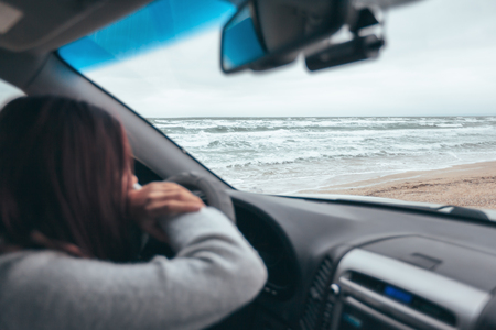 Young girl thinking in car and looking at sea waves outside. Weekend trip in bad rainy weather. Dramatic winter travel concept.の写真素材