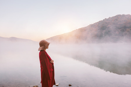 Person wrapped in warm coat outdoor standing and looking at sunrise with fog. Cold morning by the lake.の写真素材