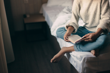 Teen girl in sweater reading a book in a hotel bedroom in the morningの写真素材