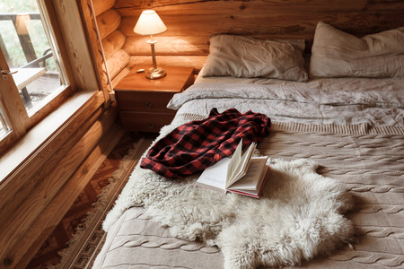 Rustic interior of log cabin bedroom with bed by big window. Opened book on sheep rug. Warm and cozy weekend morning in hotel.の写真素材
