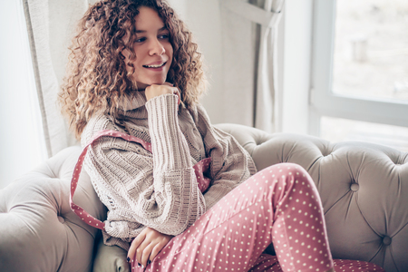 Hipster teenage girl with curly hair wearing beige knitted sweater and pink polka dot jumpsuit relaxing on a couch indoorの写真素材