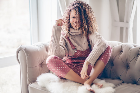 Hipster teenage girl with curly hair wearing beige knitted sweater and pink polka dot jumpsuit relaxing on a couch indoorの写真素材