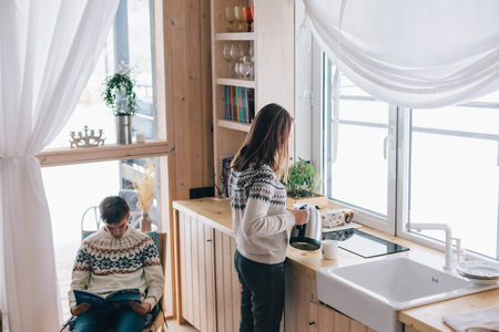 Teenage friends spending winter weekend in a cozy nordic cabin. Boy is reading and girl is making morning tea or coffee in the kitchen near big window.の写真素材