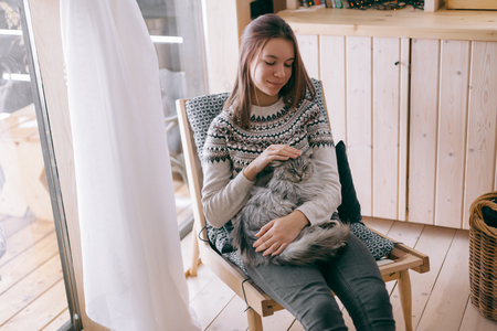 Young girl in sweater with cat relaxing on chair in log cabin near windowの写真素材