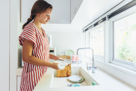 Teenage girl washing dish in the modern white kitchen. 12 years old girl doing housework.の写真素材