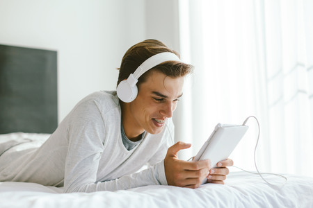 16 years old teenager using ipad and listening to music in earphones while relaxing on couch in his...