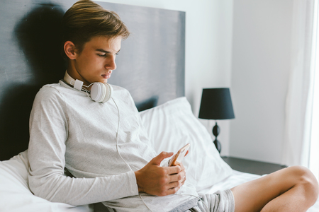 16 years old teenager using smartphone and listening to music in earphones while relaxing on couch in his roomの写真素材