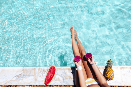 Girl sitting by pool and eating watermelon, POV photo of legs closeup. Tropical fruit diet. Summer holiday idyllic concept top view.の写真素材