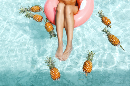 Girl relaxing on float ring in pool with pineapples in blue clear water, photo of legs closeup....