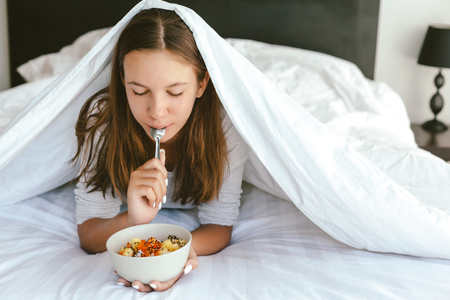 Portrait of a 12-13-14 years old teenage girl eating healthy meal in bowl while lying under blanket in bed in morningの写真素材