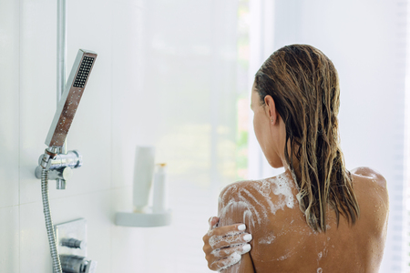 Back view of woman taking shower in modern white bathroom in the morning. Daily routine lifestyle photo.