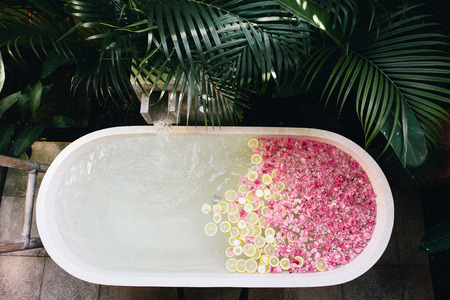 Top view of bath tub filling with water with flowers and lemon slices. Organic spa relaxation in...