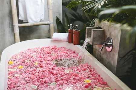 Bath tub with flower petals filling with water. Organic spa relaxation in luxury Bali outdoor bathroom.の写真素材