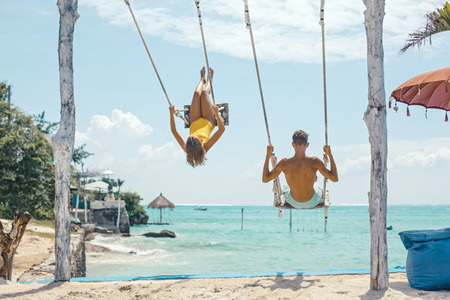 Teenage girl and boy hanging on swings with a sea view in beach cafe. Family summer vacation on paradise island.の写真素材