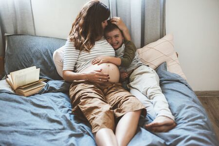Pregnant mom talking and playing with her child in the bedroom. Mother with son sharing good emotions while relaxing at home.の写真素材
