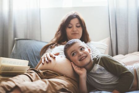 Pregnant mom talking and playing with her child in the bedroom. Mother with son sharing good emotions while relaxing at home.の写真素材