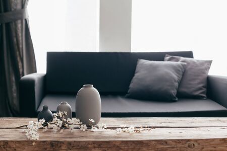 Minimalistic home decor on rustic coffee table over black sofa with cushions. Grey vases and spring flowers on wooden bench in small dark room interior. Scandinavian home style.の写真素材
