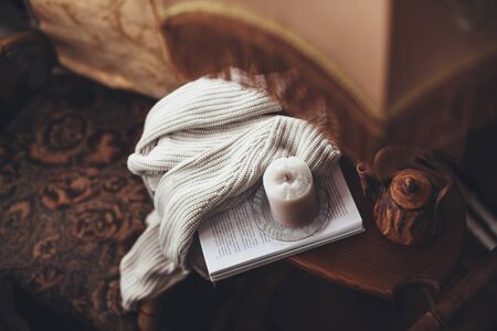 Book, candle, teapot and sweater on coffee table near armchair in wooden house. Cozy place for reading and resting at home.の写真素材