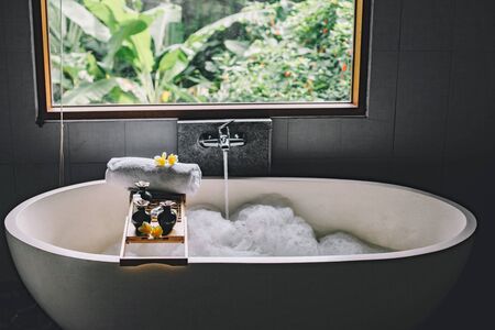 Beauty products on tray on bathtub by the window. Bath with foam is filling with water. Relaxing in Bali luxury spa hotel.の写真素材