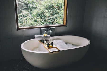 Beauty products on tray on bathtub by the window. Bath with foam is filling with water. Relaxing in Bali luxury spa hotel.の写真素材