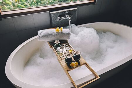 Beauty products on tray on bathtub by the window. Bath with foam is filling with water. Relaxing in Bali luxury spa hotel.の写真素材