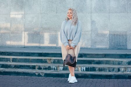 Beautiful plus size model with blond curly hair wearing grey knitted sweater and pastel skirt posing on city street. Fashion everyday outfit for cold season.の写真素材