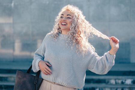 Beautiful plus size model with blond curly hair wearing grey knitted sweater and pastel skirt posing on city street. Fashion everyday outfit for cold season.の写真素材