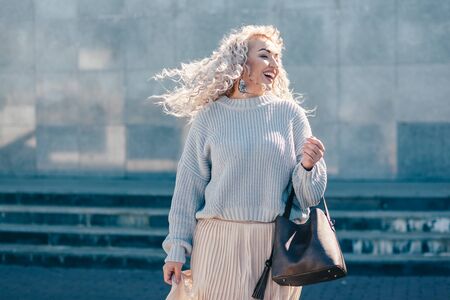 Beautiful plus size model with blond curly hair wearing grey knitted sweater and pastel skirt posing on city street. Fashion everyday outfit for cold season.の写真素材