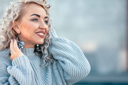 Beautiful plus size model with blond curly hair wearing grey knitted sweater and silver earrings posing on city street. Fashion everyday outfit for cold season.の写真素材