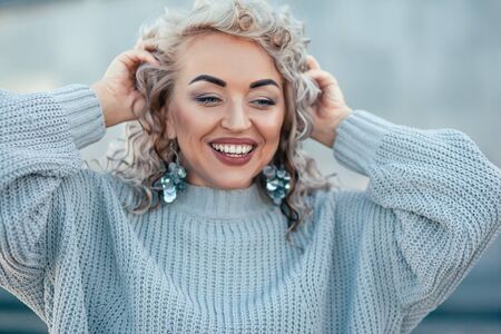 Beautiful plus size model with blond curly hair wearing grey knitted sweater and silver earrings posing on city street. Fashion everyday outfit for cold season.の写真素材