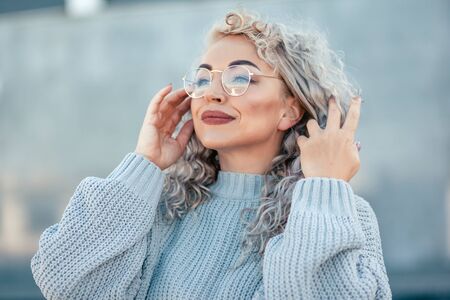 Beautiful plus size model with blond curly hair wearing grey knitted sweater and hipster glasses posing on city street. Fashion everyday outfit for cold season.の写真素材