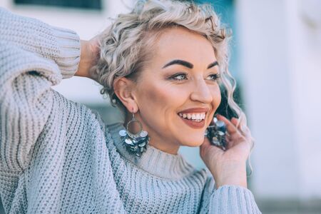 Beautiful plus size model with blond curly hair wearing grey knitted sweater and silver earrings posing on city street. Fashion everyday outfit for cold season.の写真素材