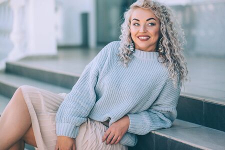 Beautiful plus size model with blond curly hair wearing grey knitted sweater and silver earrings posing on city street. Fashion everyday outfit for cold season.の写真素材