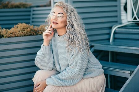 Beautiful plus size model with blond curly hair wearing grey knitted sweater and hipster glasses posing on city street. Fashion everyday outfit for cold season.の写真素材